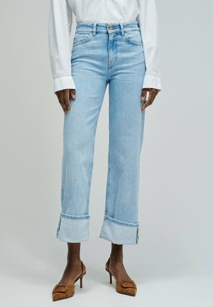 Personne portant un jean bleu clair retroussé, une chemise blanche à manches longues, et des chaussures à talons hauts pointues marron, debout sur un fond uni.