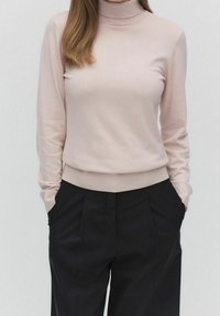 Maglione aderente rosa chiaro a collo alto con polsini e orlo a costine, abbinato a pantaloni neri a vita alta con pieghe frontali e tasche.