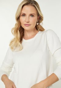 Bílý svetr s dlouhým rukávem a žebrovaným výstřihem a manžetami, s hladkou texturou. Modelka má na sobě perlové náušnice a vlnité blond vlasy.