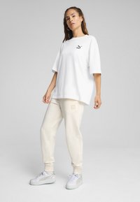 T-shirt bianca in cotone con maniche corte e logo nero, abbinata a pantaloni della tuta color crema con toppa del logo e sneakers bianche con dettagli argentati.