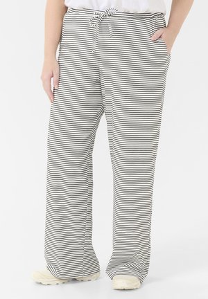 Persoon die witte en zwarte dun gestreepte wijde broek draagt met een trekkoord in de taille en witte sneakers, staand tegen een effen achtergrond.