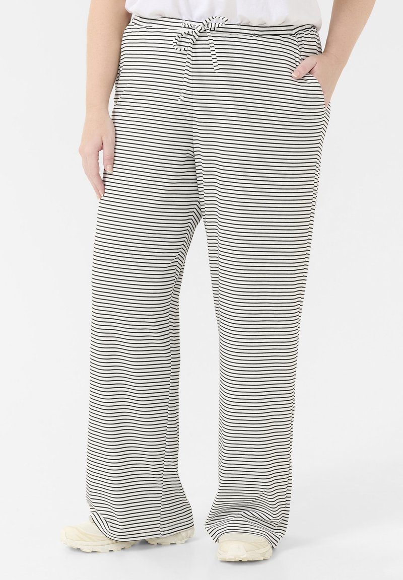 Personne portant un pantalon large à fines rayures blanches et noires avec une taille à cordon et des baskets blanches, debout devant un fond uni.