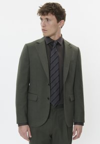 Costume vert olive à la coupe ajustée, associé à une chemise sombre et une cravate à rayures noires. Détails remarquables : revers crantés et deux boutons.