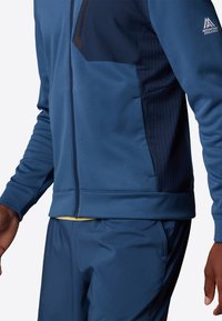 Blauwe sportjack met rits en gewatteerde marineblauwe panelen, lange mouwen en een aansluitend ontwerp, gecombineerd met bijpassende blauwe broeken.