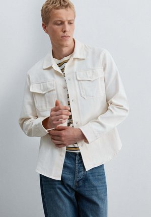 Joven con una chaqueta blanca abotonada sobre una camisa de rayas y vaqueros azules, ajustándose la manga frente a un fondo liso y claro.