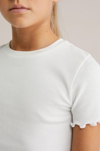 Valge ribaga T-särk, millel on ümberlõigatud kaelus ja lühikesed varrukad, millel on lainelised servad. Kangas on sileda tekstuuriga.