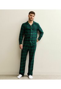 Pigiama verde a quadri realizzato in tessuto morbido, composto da una camicia con bottoni e tasca e pantaloni abbinati. Presentato su uno sfondo neutro.