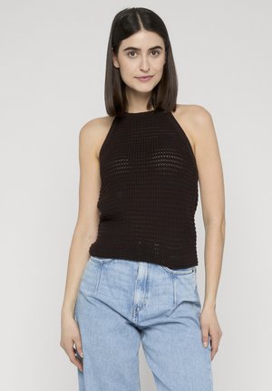 Frau mit glattem, dunklem Haar trägt ein ärmelloses, schwarzes Strick-Halftertop und hellblaue Jeans mit hoher Taille, steht vor einem schlichten hellen Hintergrund.