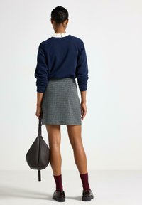 Marineblå sweatshirt med ribbede manchetter, gråternet nederdel. Holder en struktureret sort taske. Røde sokker og kraftige sorte sko. Ren baggrund.