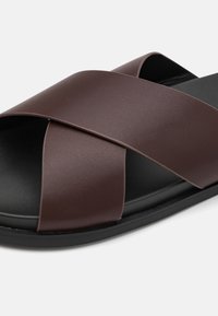 Sandale en cuir brun avec des brides croisées en matériau synthétique lisse ; semelle noire avec une finition texturée ; design minimaliste.