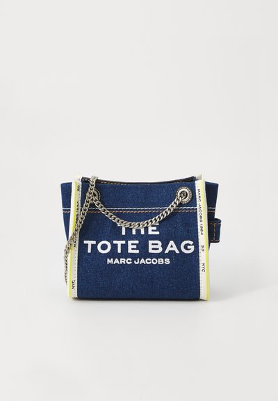 Sac fourre-tout en denim bleu Marc Jacobs avec texte blanc, anses en chaîne argentées, et bord blanc avec des détails imprimés sur les côtés.