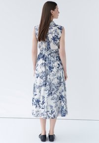 Robe maxi présentant un motif floral bleu sur fond blanc, sans manches, avec une ceinture à la taille et un tissu fluide. Des chaussures noires unies complètent la tenue.