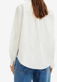 White long-sleeve shirt with a buttoned cuff, curved hem, and back yoke detail, paired with blue denim jeans showcasing a textured finish.