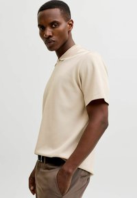 Homme aux cheveux courts et portant une boucle d'oreille, vêtu d'un polo beige à manches courtes et d'un pantalon marron, les mains dans les poches, sur un fond uni.