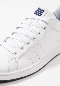 Bílé kožené tenisky s texturovaným svrškem, perforovanými detaily a plochými bílými tkaničkami. Na jazyku se nachází modrý pruhovaný logo.