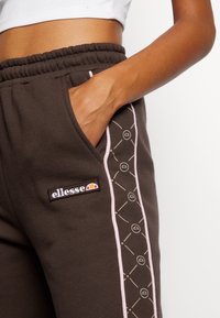 Pantaloni della tuta marrone scuro con vestibilità rilassata, caratterizzati da una striscia di accento rosa e un'etichetta con logo sulla tasca, realizzati in tessuto morbido.