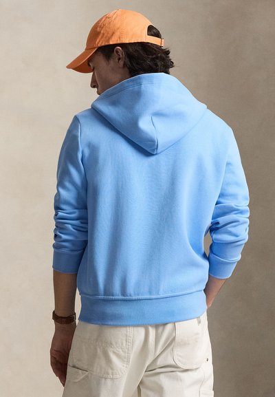 Isik, kes kannab helesinist kapuutsiga pusa ja beeže pükse, näoga eemale, oranži nokamütsi ja pruuni käekellaga neutraalsel taustal.