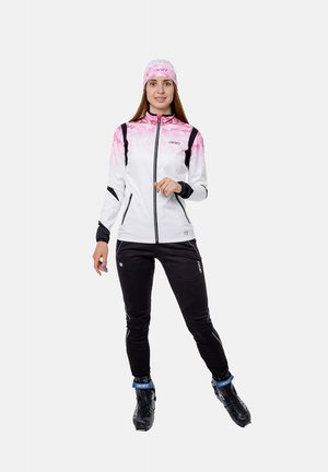 Femme portant une veste zippée rose et blanche, un bandeau assorti, un pantalon noir et des chaussures de ski noires, debout sur un fond blanc.