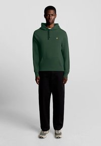 Camisola com capuz verde com um bolso frontal, cordões e um pequeno logótipo amarelo. Combinada com calças pretas largas e ténis cinzentos.