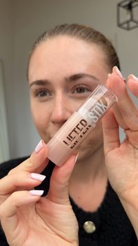 Produit à lèvres Lifter Stix dans un tube cylindrique transparent avec une teinte beige. L'étiquette présente des lettres en gras en blanc et en noir, avec une texture lisse.