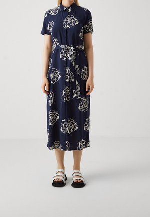 Frau trägt ein marineblaues, knöpftes Midikleid mit weißen Blumenmustern und weiße Sandalen mit schwarzen Sohlen, steht vor einem einfarbigen Hintergrund.