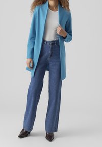 Blå ullkofta över en vit topp, kombinerad med högmidjade blå jeans och spetsiga mörka skor. Långa ärmar och öppen design fram.