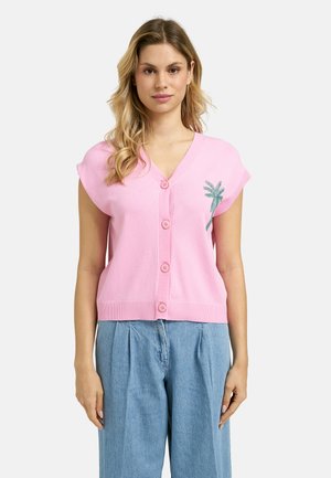 Woman wearing a pink sleeveless buttoned cardigan with a green palm tree design and high-waisted loose blue jeans, standing against a plain background.