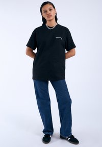 Camiseta de algodón negra de manga corta, con un pequeño logo en el pecho. Combinada con pantalones de mezclilla azul y zapatos verde oscuro.