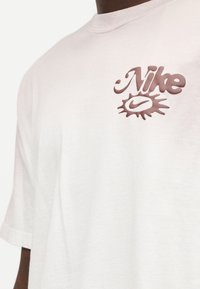 Λευκό βαμβακερό μπλουζάκι με ανάγλυφο ροζ λογότυπο που περιλαμβάνει τη λέξη "Nike" και ένα γραφικό διακοσμητικό από κάτω.