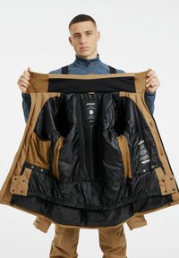 Chaqueta aislante marrón con un interior negro, múltiples bolsillos y cremalleras. El modelo la sostiene abierta para mostrar el forro y las etiquetas.