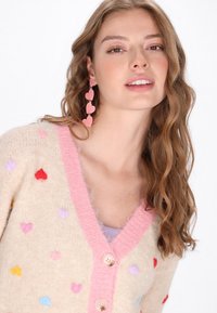 Cardigan bej tricotat cu modele colorate de inimioare, guler roz tricotat și două nasturi în față. Păr lung, ondulat, de culoare brunetă.