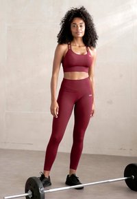 Burgunderfarget treningssett med en ribbet sports BH og høydejusterbare leggings. Utstyrt med en vektstang på gulvet, står modellen selvsikkert.