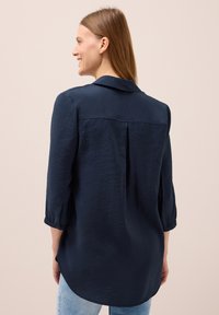 Chemise bleu marine à coupe décontractée, manches trois quarts et pli au dos. Fabriquée dans un tissu texturé, avec un col et des poignets à boutons.