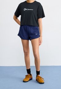 Black cropped t-shirt with white logo, paired with navy ribbed shorts featuring logo, and brown Nike sneakers with purple accents and black socks.