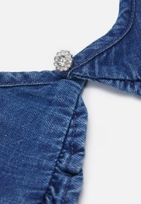 Peça em denim com uma borda franzida, apresentando um botão prateado adornado com um detalhe de gema transparente. Textura suave, cor azul profundo.