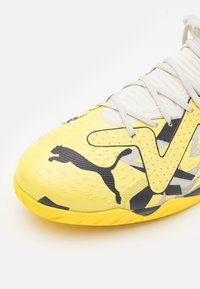 Žlutá sportovní bota s texturovaným povrchem, která má černé akcenty a geometrický vzor. Šňůrky jsou světle šedé, což podtrhuje celkový design.