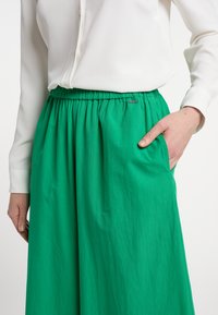 Groene, geplooide rok van lichtgewicht stof, met een elastische tailleband en zijzakken. Een klein metalen accent is zichtbaar.
