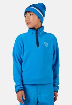Blauer Fleece-Pullover mit hohem Kragen und Reißverschluss vorne, mit einem Logo auf der Brust. Passende blaue Mütze mit Bommel.