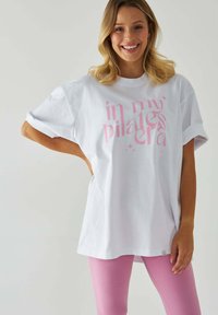 Przyduża biała bawełniana koszulka z różowym napisem "w mojej erze pilatesu", z krótkimi, podwiniętymi rękawami oraz subtelnym logo na końcu. Połączona z różowymi legginsami.