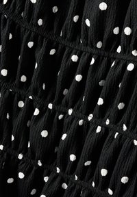 Schwarzer strukturierter Stoff mit weißen Punkte, der gefältete Bereiche und elastische Bänder aufweist und ein fließendes, geschichtetes Erscheinungsbild erzeugt.