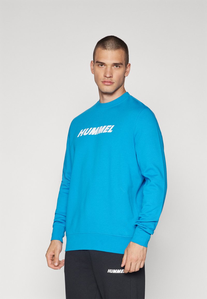 Niebieska bluza z miękkiego materiału, z okrągłym dekoltem, długimi rękawami i wyraźnym białym logo "HUMMEL" na piersi.