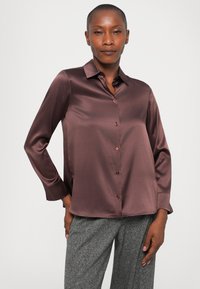 Donna che indossa una camicia button-up di satin grigio malva a maniche lunghe e pantaloni grigi con texture, in posa contro uno sfondo chiaro e semplice.
