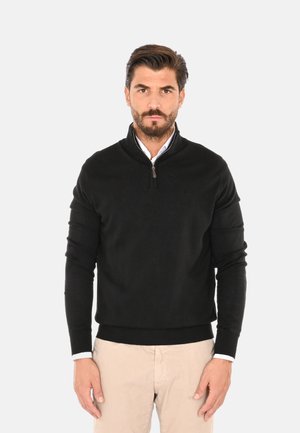 Maglione zip-up nero realizzato in tessuto morbido, con maniche lunghe e orlo a coste. Indossato sopra una camicia bianca con colletto.
