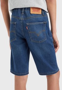 Niño que lleva pantalones cortos de mezclilla Levi's azules y una camiseta gris clara, visto desde atrás sobre un fondo sencillo.