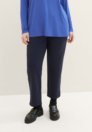 Person trägt gerade geschnittene marineblaue Hose, schwarze Socken, klobige schwarze Loafers und ein langärmliges blaues Oberteil und steht auf einem hellen Boden.