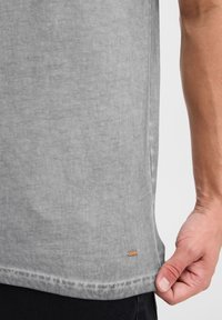 Camiseta de algodón gris con una textura suave, con un dobladillo curvado y una pequeña etiqueta de cuero cerca de la parte inferior. La mano sostiene el dobladillo.