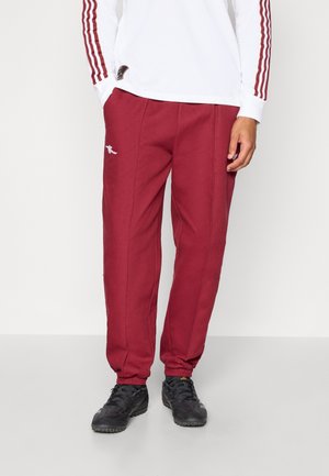 Hombre vistiendo pantalones jogger granate, zapatillas negras y una camiseta blanca de manga larga con rayas granates en las mangas, de pie contra un fondo blanco.