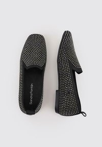 Mocasines negros sin cordones con un diseño de tachuelas, superficie texturizada y punta redonda. Tacón bajo con una pestaña trasera.