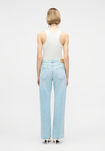 Personne aux cheveux blonds courts portant un haut blanc sans manches, un jean taille haute large bleu clair, et des chaussures blanches à talons hauts, vue de dos.