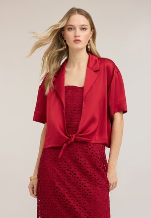 Mujer rubia con pendientes y pulsera de oro, un vestido rojo de encaje sin tirantes y una blusa roja de satén con mangas cortas y atada.